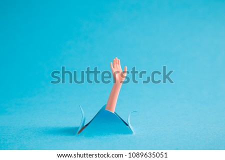 Doll arm emerging from blue paper background. Drowning minimal creative abstract concept.
