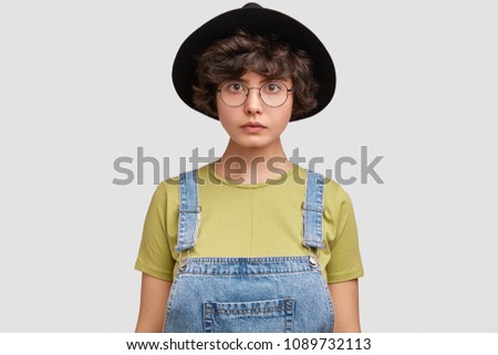 Lovely serious college student in fashionable denim overalls and hat, listens attentively information from professor, has confident look, isolated on white background. People, youth, facial expression