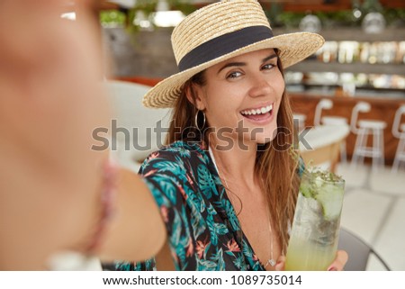 People, summer holidays and resort concept. Beautiful brunette young female with pleasant smile, wears straw hat and fashionable blouse, holds cold drink, makes selfie, enjoys good rest abroad