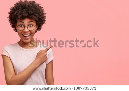 Horizontal portrait of happy dark skinned mixed race female model indicates with fore finger at blank copy space, shows place for your advertisement or promotional text. Lovely African American girl Royalty-Free Stock Photo #1089735371