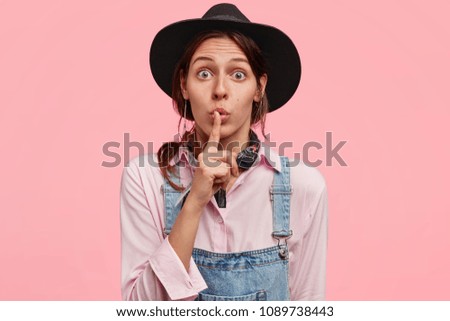 Pretty female gardener tells secret to other farmer, keep fore finger on lips, wears black hat and denim overalls, makes hush sign, looks surprisingly at camera, isolated on pink background.