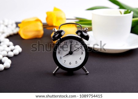 Black alarm clock near bouquet yellow tulips and cup of lemon tea on black background. Mother or Woman  Day. Greeting Card. Good Morning breakfast. Copy space. Spring. 
