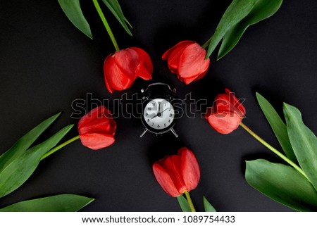 Black alarm clock near bouquet red tulips and cup of lemon tea on black background.Mother or Woman  Day. Greeting Card. Good Morning breakfast. Copy space. Spring. 
