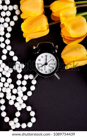 Black alarm clock near bouquet yellow tulips  on black background. Flat lay. Mother or Woman  Day. Greeting Card. Good Morning breakfast. Copy space. Spring. 
