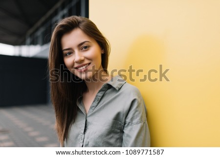 Close up portrait of young cute female wearing casual shirt posing for pictures. Beautiful smiling woman standing on the street near yellow background. Good mood.  