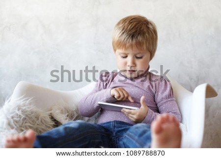 Picture of focused 5 year old girl with short hair wearing elegant violet top and jeans, absorbed in gadget, browsing websites and watching cartoons online while her mother is busy. Selective focus