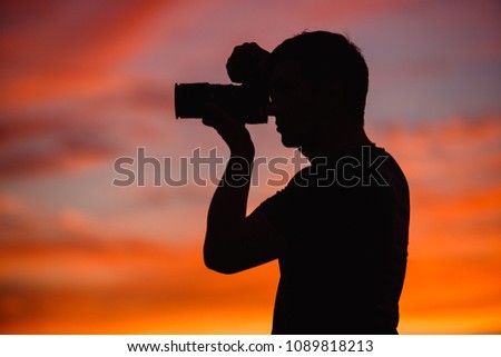 Silhouette of photographer guy with camera on sky background at sunset