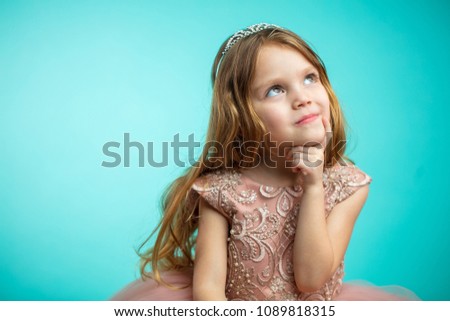 Portrait of adorable 4- year old girl with long blonde loose hair in pink princess dress looking up aside with dreamy expression isolated over blue background with copyspace for text.