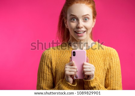 Portrait of excited cheerful cute redhaired female student showing pink smartphone isolated on pink background