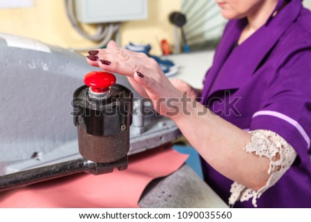 A young woman working as a seamstress in a purple unifrome presses the large red button on the industrial machine at the factory