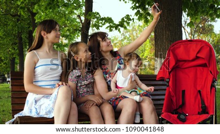Young mother is photographed with her children on phone while sitting on bench in park and smiles at little baby sitting in stroller. Family makes selfie on phone and waving.