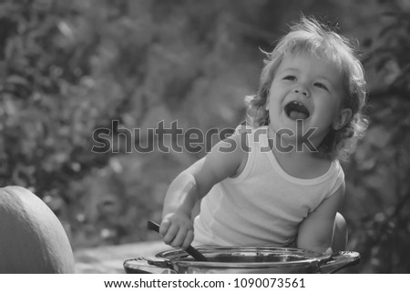 One happy smiling small child boy with blonde curly hair making food in kitchen bowl sitting at picnic with orange pumpkin sunny day outdoor on green natural background, horizontal picture