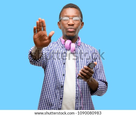 African black man student with smartphone annoyed with bad attitude making stop sign with hand, saying no, expressing security, defense or restriction, maybe pushing