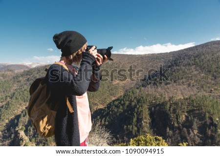 Close-up portrait of a blond girl with a digital SLR camera photographing in the mountains. Stylish freelancer photographer with backpack in mountain caucasian trip
