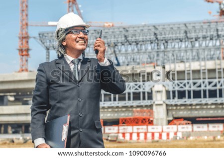 portrait of builder talking in portable radio transmitter and clipboard standing against unfinished building