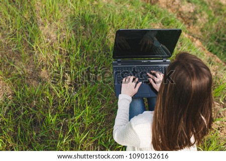 Young business woman or student sitting on grass using laptop with blank empty screen monitor in field spreading hands working outdoors on green background. Mobile Office Lifestyle concept. Back view