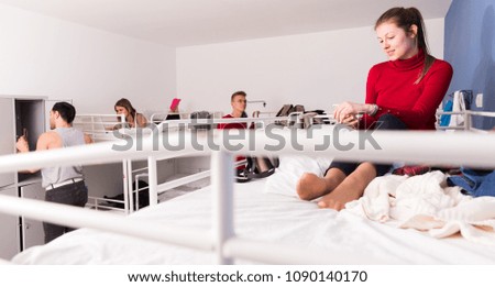 Smiling young woman sitting on top bunk of bunk bed in hostel, looking at smartphone