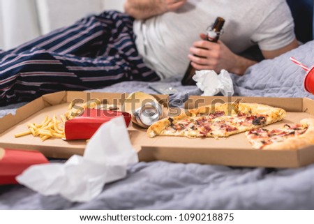 cropped image of loner eating pizza with beer on bed in bedroom
