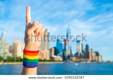 Hand wearing gay pride rainbow sweat band making 'number one' hand sign in front of city skyline