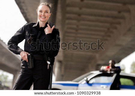 happy attractive policewoman using walkie-talkie with blurred partner near car on background Royalty-Free Stock Photo #1090329938
