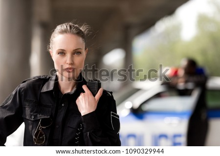policewoman using walkie-talkie and looking at camera with blurred partner near car on background Royalty-Free Stock Photo #1090329944