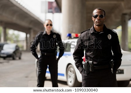police officers with hands in pockets looking at camera in front of car Royalty-Free Stock Photo #1090330040