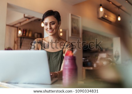 Woman entrepreneur working on laptop computer at home with a smoothie on the table. Woman with tattoos on her body working on laptop.