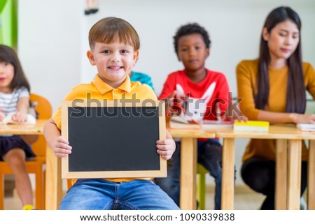 Kid boy thumbs up and holding blackboard with back to school word with diversity friends and teacher at background,Kindergarten school,mock up chalkboard for adding text.