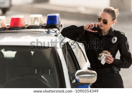 serious policewoman with burger in hand talking on radio set  Royalty-Free Stock Photo #1090354349