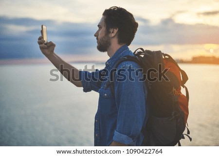 Calm young man is taking break after hiking. He is standing at the coastline and making selfie on smartphone