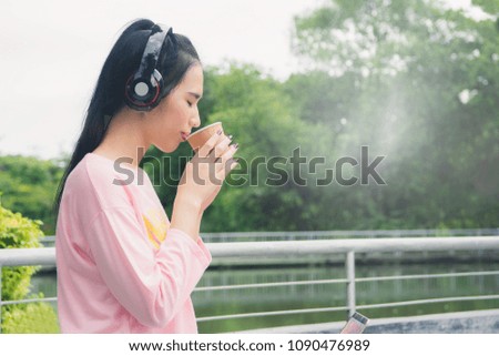 Beautiful pretty Asian woman drinking coffee with headphone in the park and outdoor public river on nature flare sun light background, coffee morning