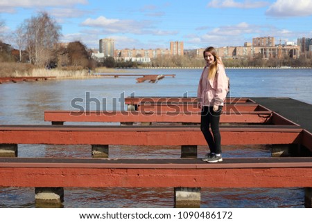 a slender blonde in a spring pink windbreaker balances on a narrow passage of the pier on the Izhevsk pond. Stunning girl with a bright sunny and spring day on the lake