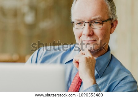 Closeup portrait of content middle-aged business man touching chin and working on laptop with blurred view in background