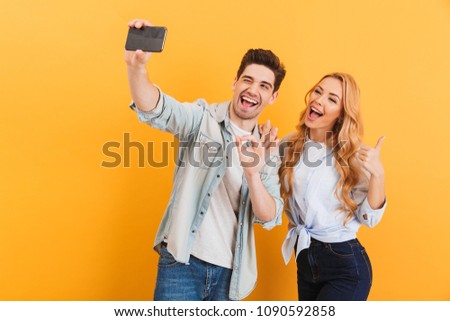 Portrait of young man and woman taking selfie photo on mobile phone while gesturing at camera with fingers isolated over yellow background