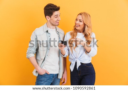 Photo of happy people man and woman laughing while showing at cell phone isolated over yellow background