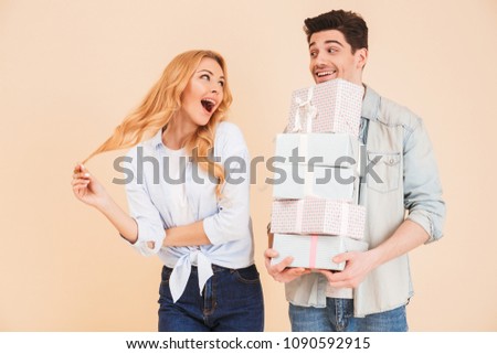 Image of charming woman rejoicing and expressing surprise while handsome man holding lots of gift boxes isolated over beige background