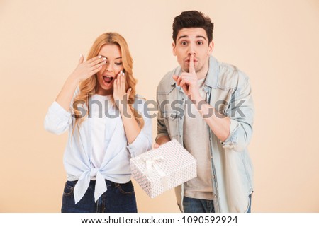 Image of young man with present box keeping finger at lips and asking to keep secret while standing near woman peeking at gift isolated over beige background