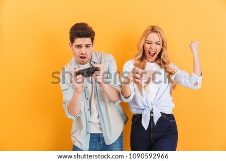 Image of excited young man and woman playing together and competing in video games on smartphones isolated over yellow background Royalty-Free Stock Photo #1090592966