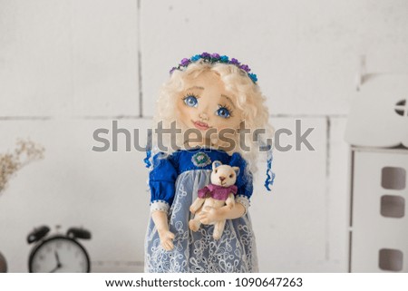 Portrait of textile handmade vintage doll with blue eyes, long blond hair in old blue textile dress with gentle print, in white shirt with purple boots on white background with toy house. She holding