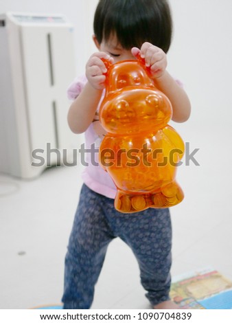 Little Asian baby girl, 13 months old, lifting up and carrying a heavy piggy bank filled with many coins - big and small muscles development in children 