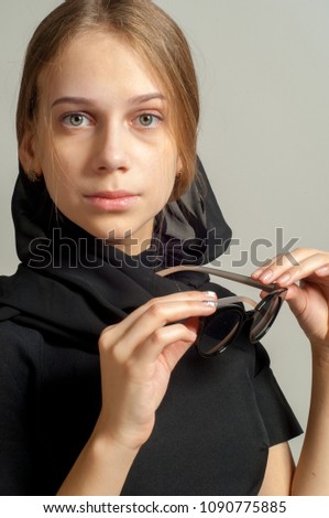 A 14-year-old girl was photographed in the studio. The girl wears a black dress. Sunglasses. And a black headscarf dressed on his head.