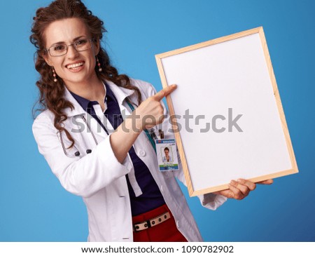 smiling physician woman in white medical robe pointing at blank board against blue background