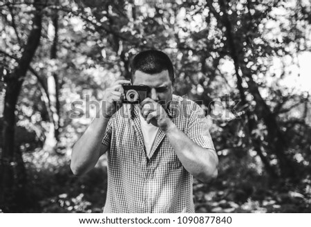 Young guy in checkered shirt is standing in the green forest with an old vintage photo camera. Hipster taking photos outdoors. Lifestyle travelling shot. Black and white photo