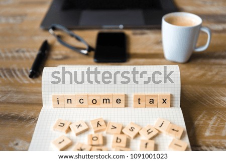 Closeup on notebook over wood table background, focus on wooden blocks with letters making Income Tax words. Business concept image. Laptop, glasses, pen and mobile phone in a defocused background.