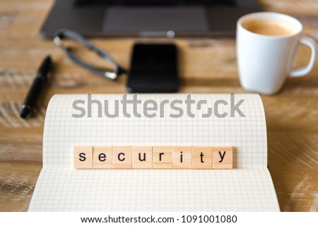 Closeup on notebook over wood table background, focus on wooden blocks with letters making Security word. Business concept image. Laptop, glasses, pen and mobile phone in a defocused background.