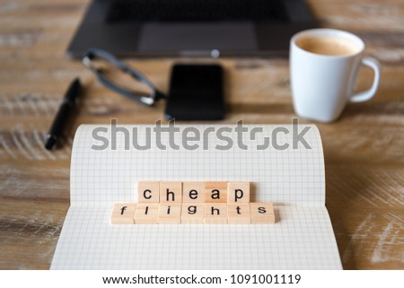 Closeup on notebook over wood table background, focus on wooden blocks with letters making Cheap Flights words. Business concept image. Laptop, glasses, pen and mobile phone in a defocused background.