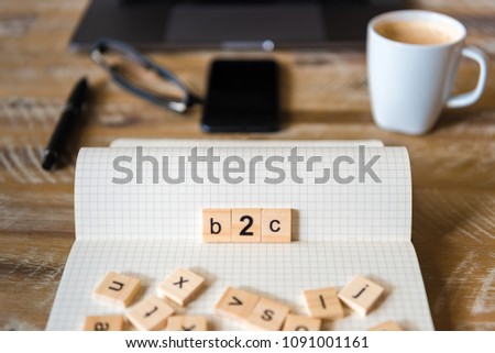 Closeup on notebook over wood table background, focus on wooden blocks with letters making B2C word. Business concept image. Laptop, glasses, pen and mobile phone in a defocused background.