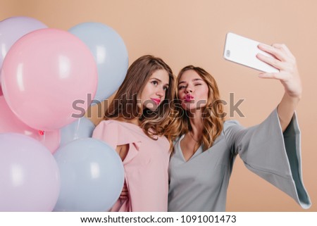 Shy girl with pink lipstick posing with party balloons. Emotional blonde woman using phone for selfie and making kissing face expression.