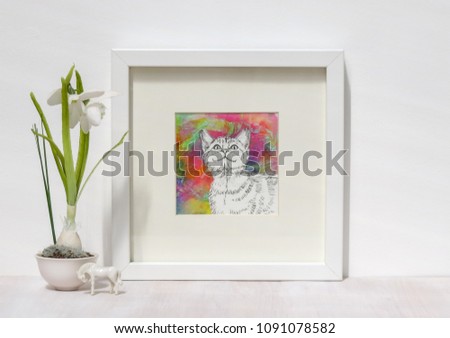 White interior display. Drawing of smiling cat on a vibtant colourful collaged background in a frame. With a snowdrop on a shelf.