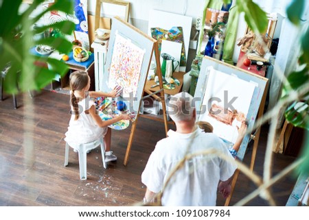 High angle portrait of children painting on easels during art class and teacher watching them in cozy studio decorated with plants